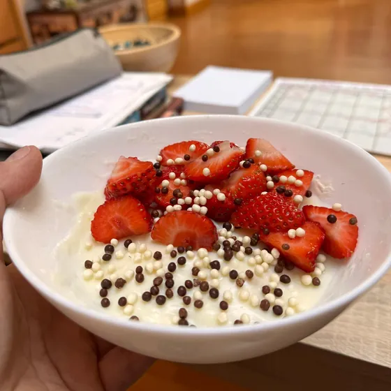 Un bol de yogur con fresas y cereales sujeto con la mano en un escritorio, entre papeles